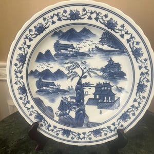 Könnte beinhalten: Ein weißer Keramikteller mit blauem Landschaftsdesign. Der Teller hat einen gewellten Rand mit Blumen- und Blättermustern. Das zentrale Design zeigt Berge, Gebäude und Bäume in einem klassischen Blau-Weiß-Farbschema.