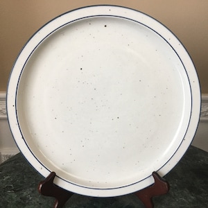 Puede incluir: Un plato de servir blanco de cerámica con un borde azul. El plato tiene un acabado moteado y es perfecto para servir aperitivos, postres u otros platos.