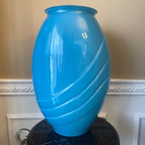 以下が含まれることがあります： 滑らかで光沢のある仕上げの大きな薄い青色のセラミック花瓶。花瓶は丸みを帯びた楕円形で、隆起した水平線が渦巻き模様を作り出しています。