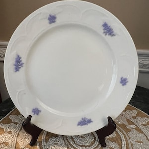 Puede incluir: Un plato de cerámica blanco con borde festoneado y detalles florales lavanda. El plato se exhibe sobre un soporte marrón oscuro, mostrando su diseño. Este plato decorativo es adecuado para servir alimentos.