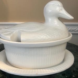 Op de afbeelding: Witte keramische serveerschaal in de vorm van een eend, met een deksel en een bijpassende voet. De schaal heeft een geribbeld ontwerp en een gladde, glanzende afwerking.