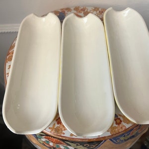 May include: Three white ceramic serving dishes with a rounded, elongated shape. The dishes are arranged on a decorative surface with a floral pattern.