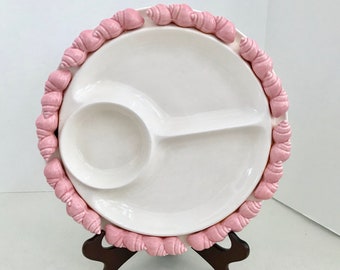 Può includere: Un piatto da portata in ceramica bianca con tre sezioni, decorato con un bordo di conchiglie rosa. Il piatto è su un supporto in legno marrone.