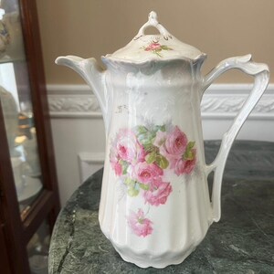 Puede incluir: Una cafetera de porcelana blanca con textura acanalada vertical y un delicado patrón floral de rosas rosadas con hojas verdes. La cafetera incluye una tapa a juego con un pequeño remate, un asa curva y un borde de pico festoneado.
