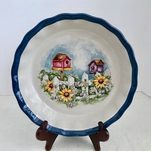 May include: A white pie plate with a blue rim and a whimsical design of birdhouses, sunflowers, and a white picket fence.