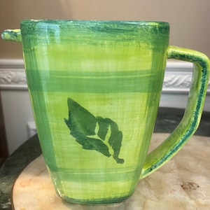 May include: Green ceramic pitcher with a leaf design and a checkered pattern. The pitcher has a handle and a spout.