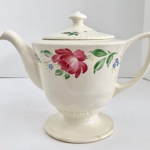 May include: A white ceramic teapot with a floral design. The teapot has a lid with a small hole in the top and a base with a pedestal. The floral design features pink roses, green leaves, and blue flowers.