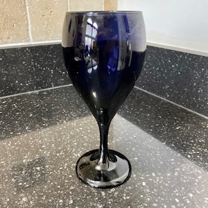 Puede incluir: Una sola copa de vino azul oscuro, elegante. La copa tiene un tallo largo y un cuenco ancho, que refleja la luz. Está hecha de un material oscuro y transparente, con una base redonda. La copa está sobre una superficie moteada.