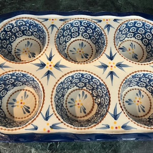 Puede incluir: Molde de cerámica para muffins con seis huecos individuales. El molde presenta un diseño floral azul y blanco con un borde festoneado y un borde azul oscuro. El interior de cada hueco tiene un patrón floral similar.