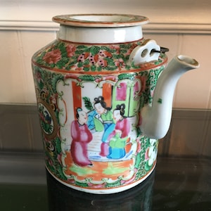 Puede incluir: Una tetera de porcelana blanca con tapa y pico. La tetera está decorada con un patrón floral colorido y una escena de personas en un jardín. La tetera mide aproximadamente 18 cm de alto.