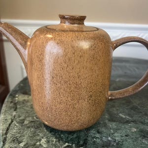 May include: A ceramic teapot with a speckled tan and brown glaze. The teapot features a cylindrical body, a curved handle, a pointed spout, and a matching lid with a small knob. The finish has a textured appearance with darker brown flecks.