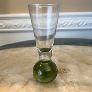 May include: A clear glass shot glass with a green glass base. The glass is tall and narrow, with a rounded bottom.