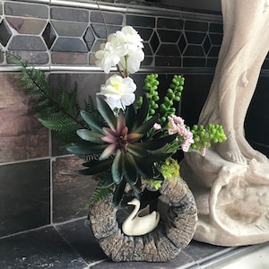 Puede incluir: Una figurita de cisne de cerámica blanca se encuentra en una maceta de madera rústica con una planta suculenta, flores blancas y follaje verde.
