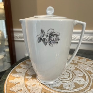 Op de afbeelding: Witte keramische koffiepot met een deksel en handvat. Een grijs en wit rozenontwerp staat op de voorkant. De pot staat op een ronde, decoratieve, beige en gouden kralen placemat.