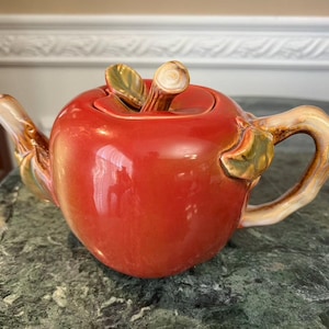 May include: A ceramic teapot shaped like a red apple with a glossy finish. The handle and spout are tan, and the lid features a stem and green leaves. The teapot is designed to resemble a realistic apple.