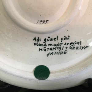 May include: White ceramic plate with a hand-painted design. The back of the plate has the text "Adi g&uuml;zel &ccedil;ini Hand made special K&uuml;tahya T&uuml;rkiye Fahriye" and the year "1993".