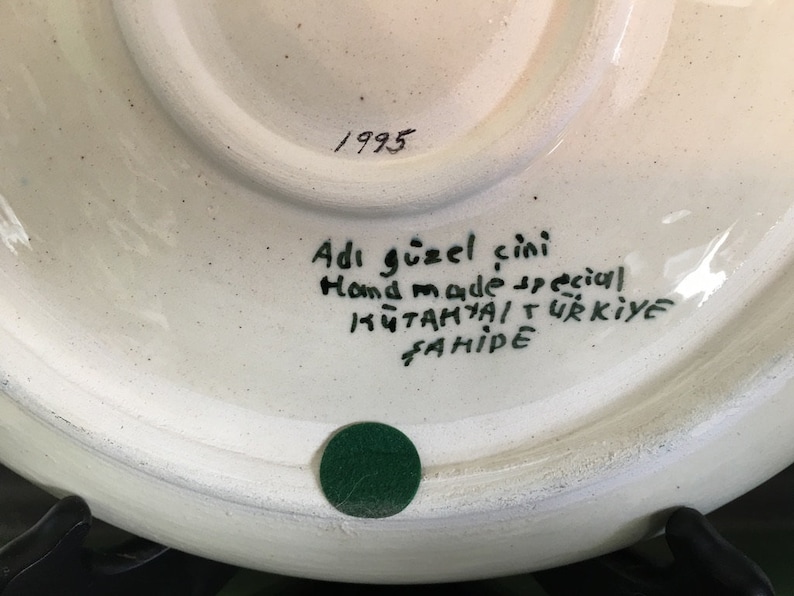 May include: White ceramic plate with a hand-painted design. The back of the plate has the text "Adi g&uuml;zel &ccedil;ini Hand made special K&uuml;tahya T&uuml;rkiye Fahriye" and the year "1993".