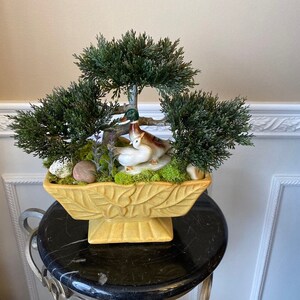 May include: A yellow ceramic planter with a leafy design features a miniature artificial bonsai tree with two white ceramic birds. The planter is sitting on a black pedestal.