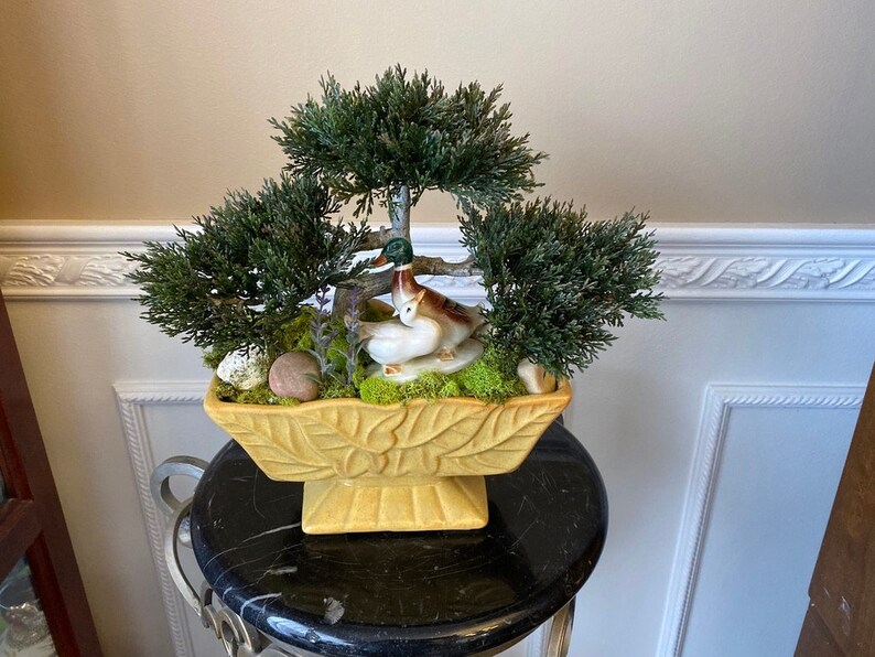 May include: A yellow ceramic planter with a leafy design features a miniature artificial bonsai tree with two white ceramic birds. The planter is sitting on a black pedestal.