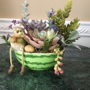 Può includere: Un vaso verde a forma di anguria con una piccola statuetta di uccello seduta sul bordo. Il vaso è pieno di piante grasse e altre piante verdi.