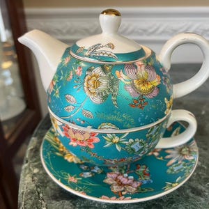 Op de afbeelding: Een turquoise theeservies met een bloemenpatroon. De set bestaat uit een theepot, een kopje en een schotel. De theepot heeft een witte tuit en handvat, en een goudkleurige knop op het deksel. Het bloemenpatroon heeft roze, gele en witte bloemen.