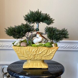 May include: A yellow ceramic planter with a leaf design, filled with moss, small rocks, and artificial greenery. Two ceramic ducks are nestled in the greenery.