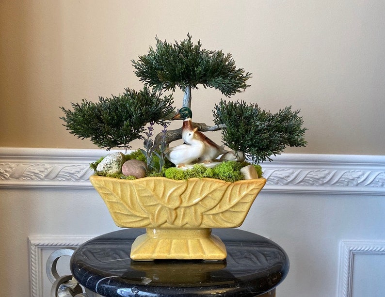 May include: A yellow ceramic planter with a leaf design, filled with moss, small rocks, and artificial greenery. Two ceramic ducks are nestled in the greenery.