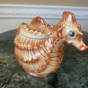 May include: A ceramic seahorse-shaped lidded container with a glossy, iridescent orange and cream finish. The body features textured, shell-like ridges and a prominent dorsal fin. The seahorse head has a distinct snout and a single visible eye.
