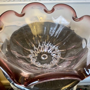 May include: A clear glass bowl with a ruffled edge and a purple tint. The bowl is made of thick glass and has a delicate, feminine design.