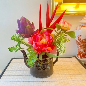 Puede incluir: Una tetera negra con diseño floral está llena de un arreglo colorido de flores artificiales, incluyendo una alcachofa morada, peonías rosas y hojas tropicales rojas.