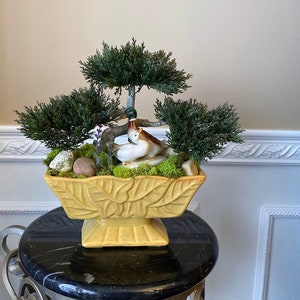 May include: A yellow ceramic planter with a leaf design, featuring a miniature bonsai tree with green foliage and two ceramic birds. The planter is sitting on a black pedestal.