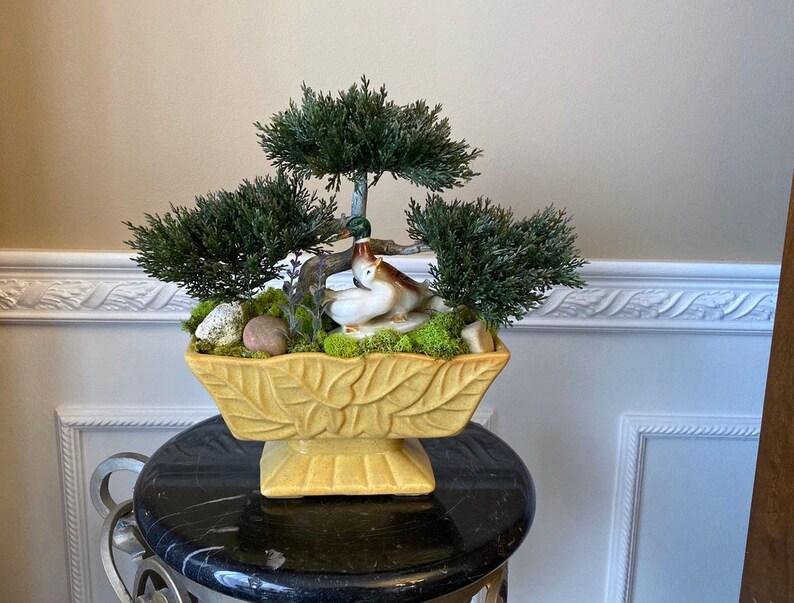 May include: A yellow ceramic planter with a leaf design, featuring a miniature bonsai tree with green foliage and two ceramic birds. The planter is sitting on a black pedestal.