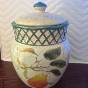 Puede incluir: Un tarro de cerámica con tapa, con una base color crema con acabado agrietado. El tarro está adornado con hojas verdes y un diseño de fruta amarilla y naranja. El borde tiene un patrón de celosía verde.