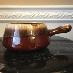 Puede incluir: Un plato para servir de cerámica esmaltada marrón y blanco con un solo asa. El plato tiene una forma redondeada y un acabado liso y brillante.