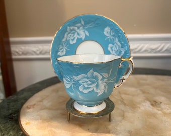 Può includere: Un set da tè composto da una tazza e un piattino di colore azzurro chiaro con un motivo floreale bianco. Il set ha un bordo dorato.