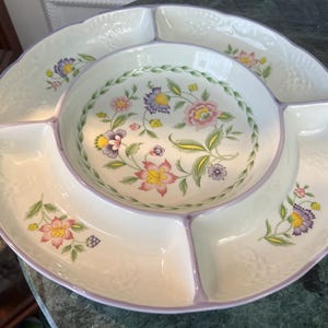 May include: A white divided serving dish with a floral design. The dish has a central bowl and five surrounding compartments. The floral pattern includes pink, yellow, and purple flowers with green leaves. The rim is trimmed with a lavender color.