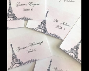 Puede incluir: Conjunto de seis tarjetas de lugar con una ilustración en blanco y negro de la Torre Eiffel. Cada tarjeta tiene un fondo rosa e incluye un nombre y el texto "Mesa 6".