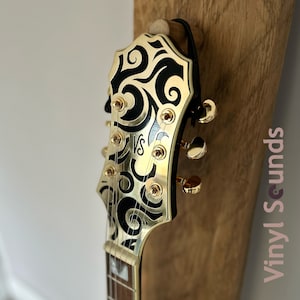 May include: Close-up of a gold electric guitar headstock with black and gold swirling designs. The headstock has gold tuning pegs and the text "VS" is visible on the face.