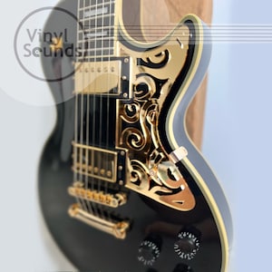 Puede incluir: Guitarra eléctrica negra con herrajes dorados y un intrincado diseño de filigrana dorada en el cuerpo. La guitarra tiene una golpeador negro y un solo corte.