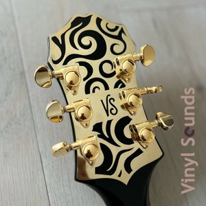 May include: Close-up of a black electric guitar headstock with gold hardware and a black and white swirl design. The headstock features six gold tuning keys with the word "Grover" printed on each key. The headstock also features the word "VS" printed in gold letters.