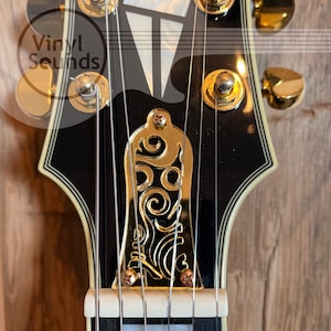 Puede incluir: Primer plano de la pala de una guitarra eléctrica negra y dorada. La guitarra tiene clavijas de afinación doradas y una tapa de varilla de armadura dorada con un diseño intrincado. La guitarra tiene seis cuerdas.