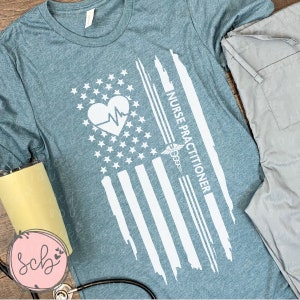 May include: Teal blue t-shirt with a white distressed American flag design. The flag has a heart with a heartbeat line inside and the text "NURSE PRACTITIONER" on the right side.