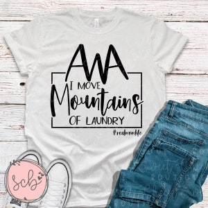 May include: White t-shirt with the text "AMA I MOVE Mountains OF LAUNDRY #realmomlife" in black. The shirt is paired with blue jeans and white sneakers on a light wood background.