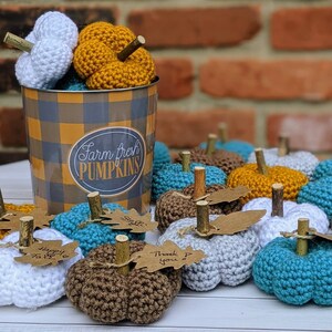 May include: A collection of crocheted pumpkin decorations in various colors, including teal, brown, white, and gray. The pumpkins are arranged on a white surface with a brown and white checkered container in the background. The container has the text "Farm Fresh Pumpkins" printed on it.