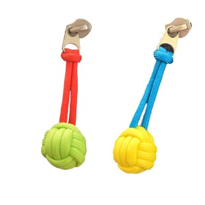 May include: Two zipper pulls with colorful accents. One features a neon green knot and red cord, the other a yellow knot and blue cord. Both have gold-colored metal zipper attachments.