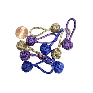 May include: A collection of seven zipper pulls made from woven cord, featuring spherical monkey fist knots in shades of gold, purple, and blue. Each pull has a looped cord end. A copper-colored U.S. penny is included for scale reference.