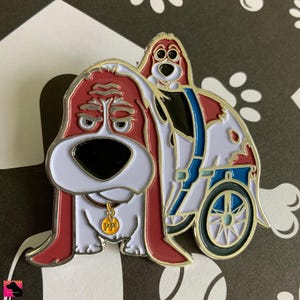 以下が含まれることがあります： 車椅子に乗った漫画の犬と、その背中に乗っている別の犬を描いた赤と白のエナメルピン。車椅子の犬は、"Pops"と書かれたタグが付いた金の首輪をしています。