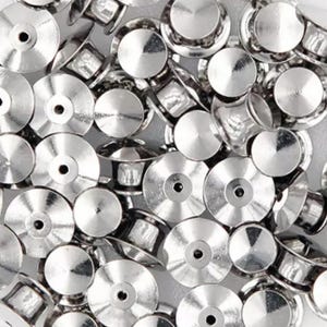 May include: A close-up of a pile of silver-toned metal pin backs. The pin backs are round and have a small hole in the center. They are used to attach pins, badges, and other items to clothing or fabric.