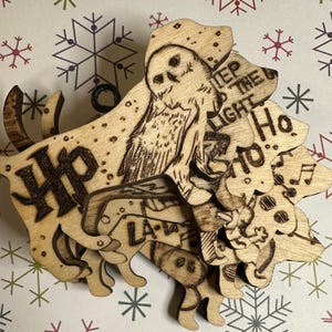 May include: Wooden Christmas ornament featuring a layered design with a Harry Potter theme. The ornament includes an owl, the letters "HP", and the words "Keep the Light Ho Ho Ho". The ornament is made of light-colored wood.