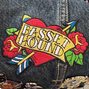 Könnte beinhalten: Dunkelblaue Jeans-Baseballkappe mit einem Aufnäher. Der Aufnäher zeigt ein rotes Herz, Rosen und einen Pfeil mit dem Schriftzug "BASSET HOUND" auf einem gelben Banner. Die Kappe hat Metallnieten.